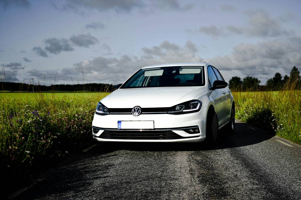 Weisser VW Golf, schräg von vorne, auf einer Landstrasse zwischen Wiesen