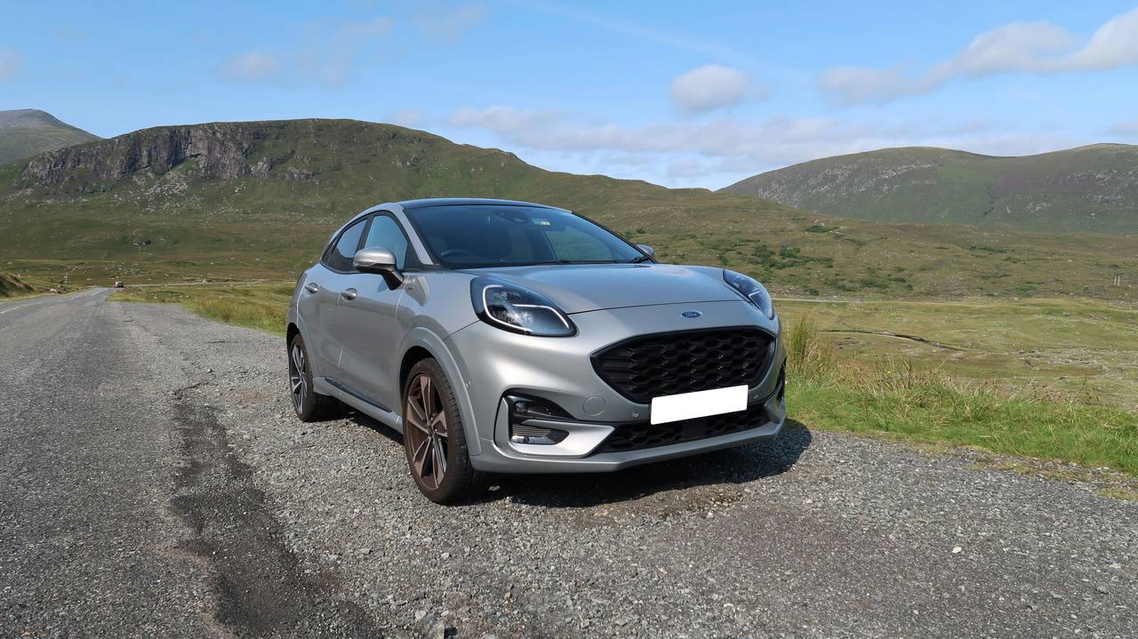 Silberner Ford Puma, schräg von vorne, auf einem Kiesplatz vor grünen Hügeln