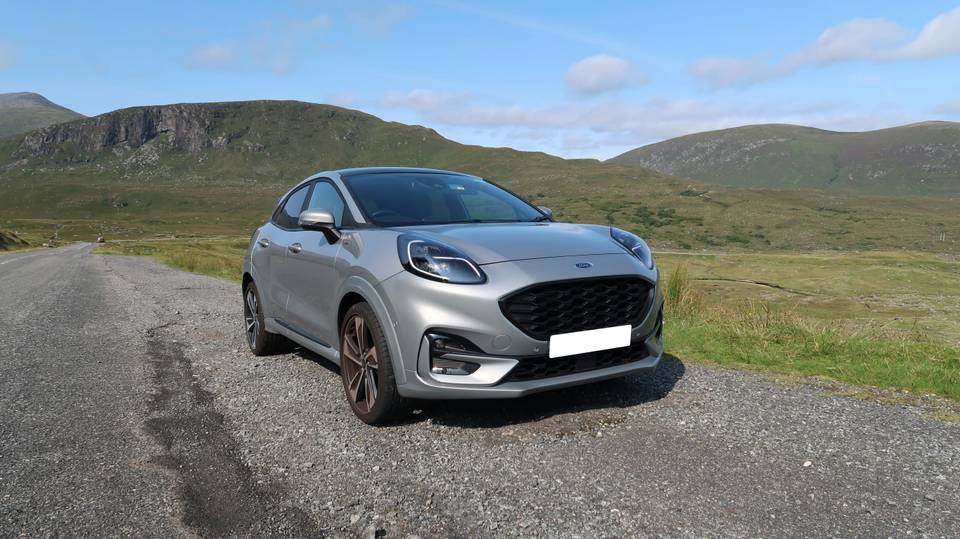 Silberner Ford Puma, schräg von vorne, auf einem Kiesplatz vor grünen Hügeln